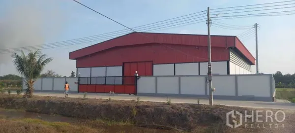Warehouse for rent or sale in Khlong Luang, Pathum Thani - Side elevation of the red steel warehouse and perimeter wall facing the paved road with utility poles.