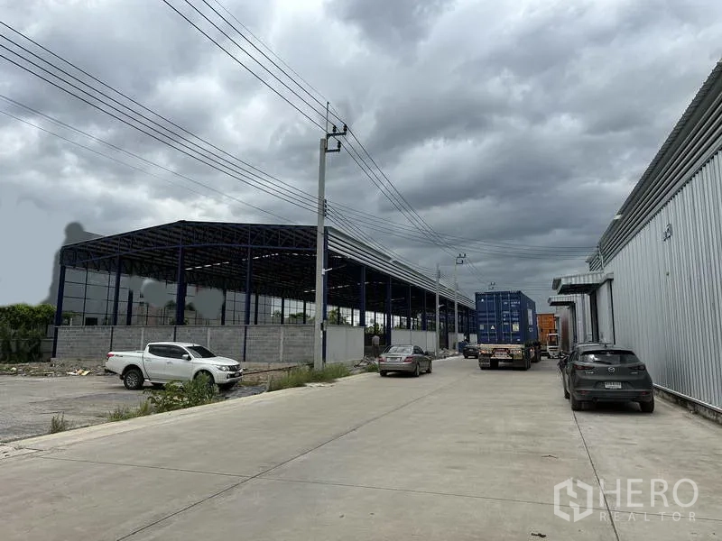 货栈 出租在Lat Krabang, Bangkok - 工业道路景观，仓库旁有集装箱卡车，邻侧钢结构雨棚在建。