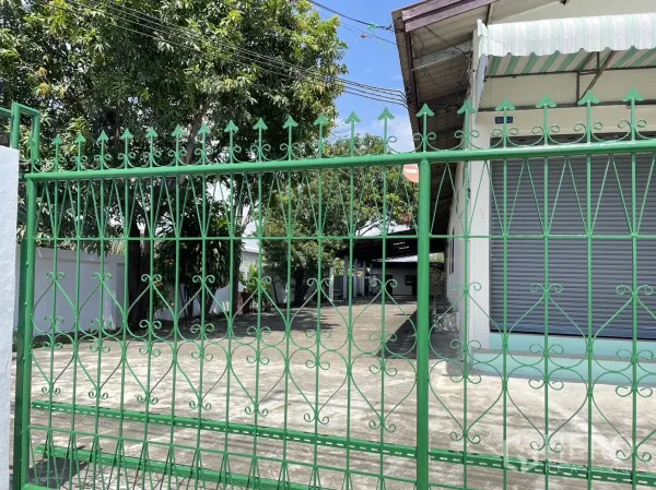 Factory for rent or sale in Pathum Thani - Ornate green gate opening to a concrete driveway with a white factory and roller shutters behind trees.