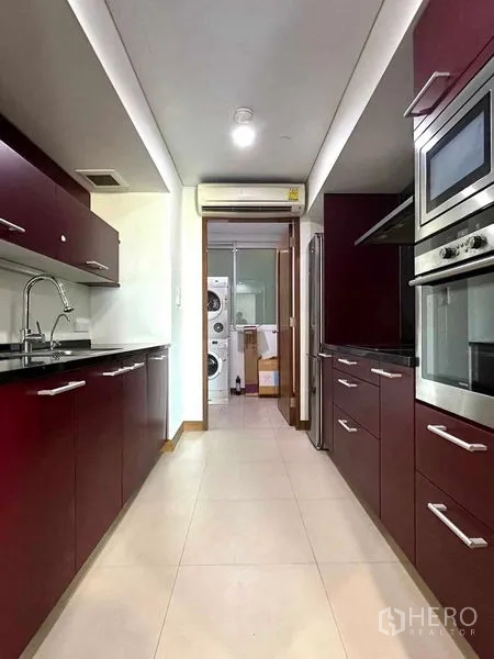 公寓 出租在Pathum Wan, Bangkok - 走廊式厨房，酒红色橱柜，内置烤箱与不锈钢冰箱，通向洗衣区。