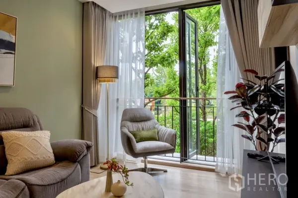 Condominium for rent or sale in Watthana, Bangkok - Bright living room with armchairs opening to a balcony overlooking lush trees.