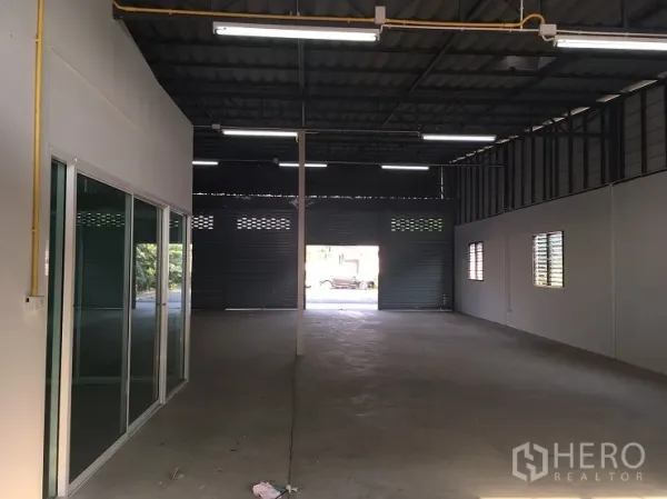 Warehouse for rent in Lam Luk Ka, Pathum Thani - Open-plan warehouse interior with high steel roof, bright fluorescent lighting, concrete floor and rear roller shutter access.