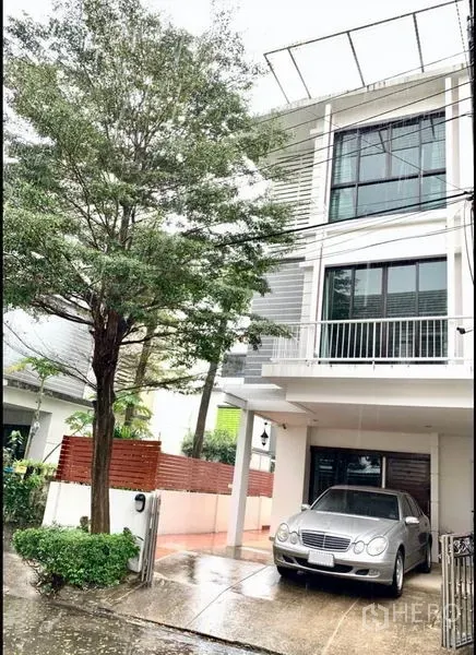 Townhouse for sale in Khlong Toei, Bangkok - Modern three-storey townhouse with balcony, large windows and a covered carport on a tree-lined lane.