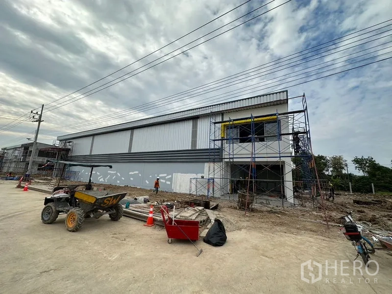 Warehouse for rent or sale in Lam Luk Ka, Pathum Thani - Side elevation of a new steel‑clad warehouse with scaffolding and tools along the roadside.