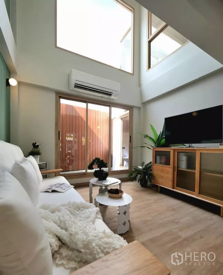Townhouse for rent in Watthana, Bangkok - Bright living room with tall windows and sliding doors to a patio, white sofa and wooden media cabinet.