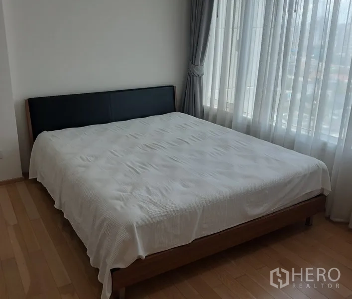 Condominium for rent in Khlong Toei, Bangkok - Corner bedroom with large windows, double bed, grey curtains, and city outlook.