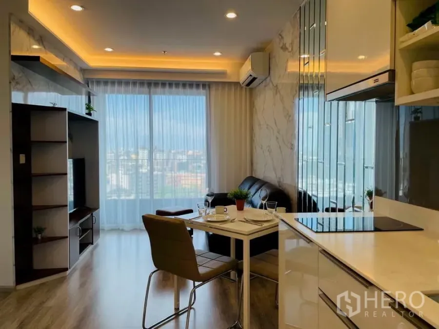 Condominium for rent in Bang Na, Bangkok - Open-plan living room with kitchenette, dining table and floor-to-ceiling windows offering a city view.