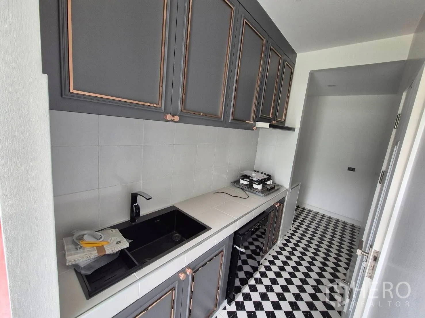 Detached House for sale in Bang Kruai, Nonthaburi - Galley kitchen with grey cabinets, black sink, gas hob and checkered floor tiles.