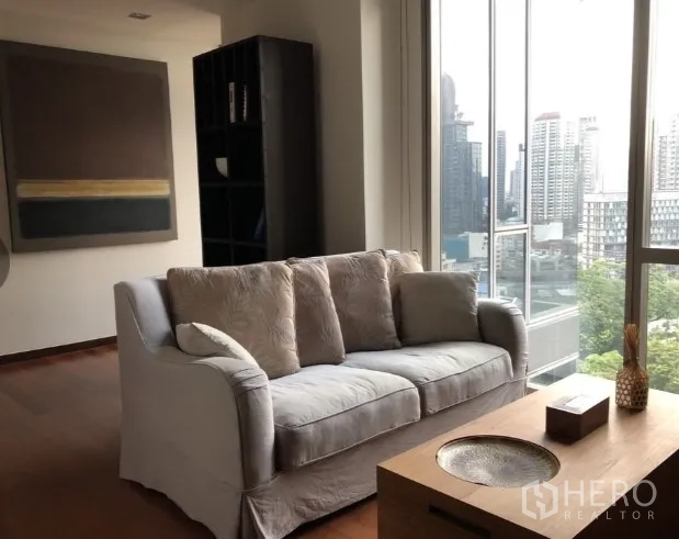 Condominium for rent or sale in Khlong Toei, Bangkok - Bright living room with cushioned sofa, wood floors, and large windows framing a Bangkok skyline view.