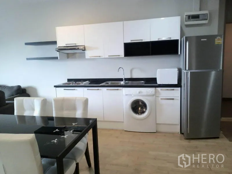 Condominium for rent or sale in Watthana, Bangkok - Modern kitchen in an Ekkamai condo featuring white cabinetry, black countertop, stainless refrigerator, in-unit washing machine, microwave, and adjacent dining area.