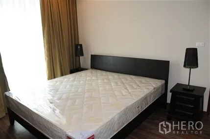 Condominium for rent in Khlong Toei, Bangkok - Bedroom with large bedframe, bedside tables and lamps beside curtained window.