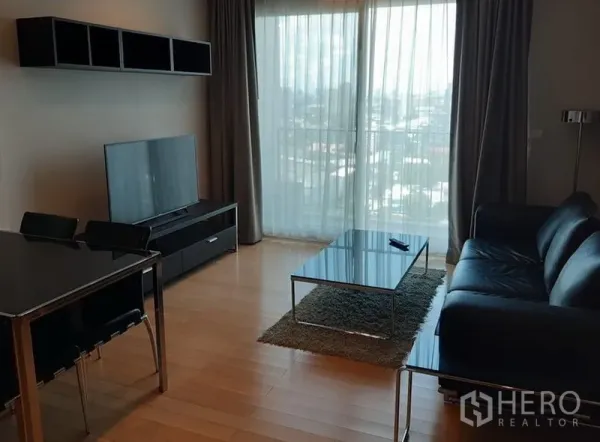 Condominium for rent in Khlong Toei, Bangkok - Bright living room with black leather sofa, TV console, glass coffee table and floor‑to‑ceiling window with city view.