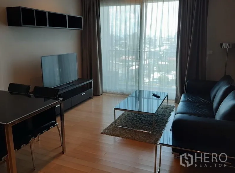 Condominium for rent in Khlong Toei, Bangkok - Bright living room with black leather sofa, TV console, glass coffee table and floor‑to‑ceiling window with city view.