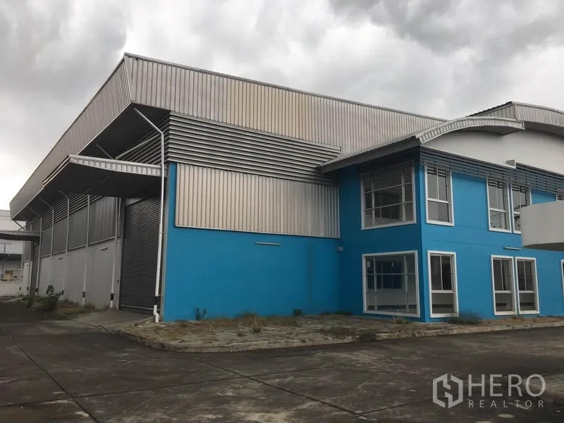 Factory for rent in Samut Prakan - Modern blue factory exterior with attached two-storey office, metal cladding and wide concrete yard under cloudy sky.