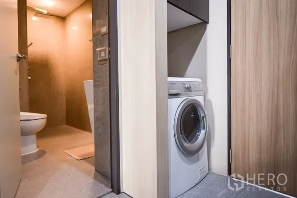 Condominium for rent in Khlong Toei, Bangkok - Laundry closet with a front-loading washing machine beside a modern bathroom with a rain shower.