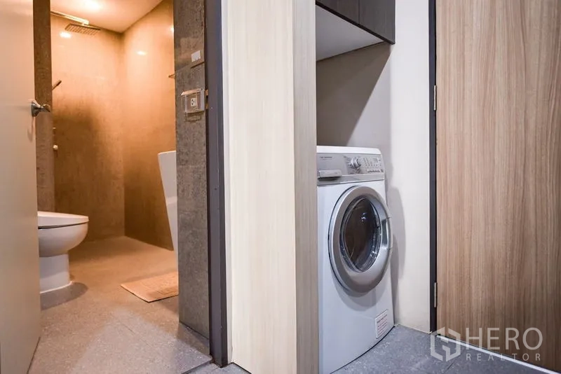 Condominium for rent in Khlong Toei, Bangkok - Laundry closet with a front-loading washing machine beside a modern bathroom with a rain shower.