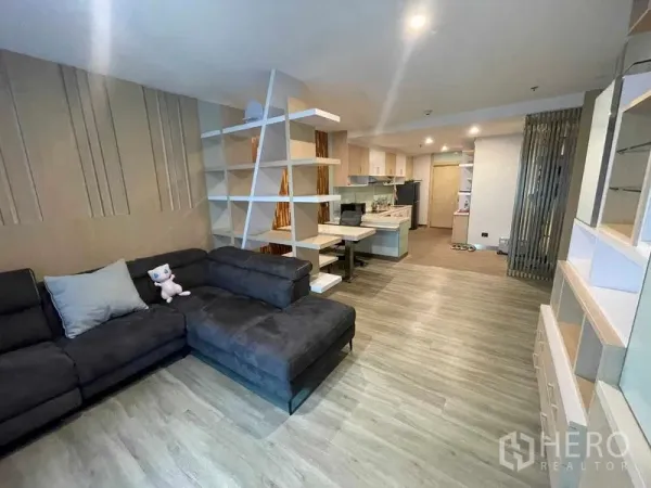 Condominium for rent in Sathon, Bangkok - Open-plan living room with L-shaped charcoal sofa, feature shelving divider and wood floors at Natural Place Suite.