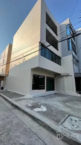 店屋 出租在Bang Kapi, Bangkok - 转角商铺外立面，白色墙面、大面积玻璃与门前停车位。