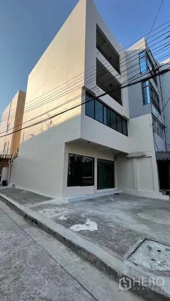 店屋 出租在Bang Kapi, Bangkok - 转角商铺外立面，白色墙面、大面积玻璃与门前停车位。