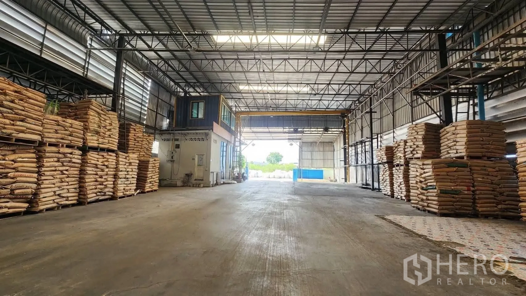 仓库 for rent in Bang Phli, Samut Prakan - 高净空仓库内部，钢桁架与采光板，上下堆放物料，旁有带窗办公室和宽敞出入口。