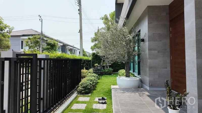 บ้าน ให้เช่าหรือขายในBang Na, Bangkok - เฉลียงหน้าบ้านและสนามหญ้าจัดแต่ง พร้อมต้นไม้ในกระถางข้างรั้วเหล็ก