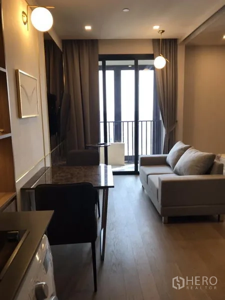 公寓 出租在Watthana, Bangkok - 现代公寓客厅，灰色沙发、餐桌、小厨房、洗衣机与通往阳台的落地门，位于曼谷瓦塔那区。