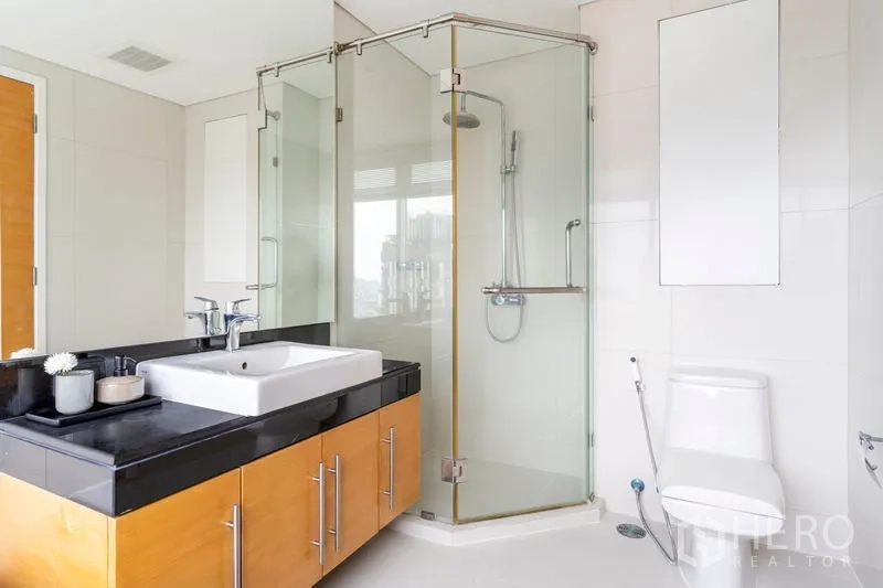 Condominium for rent in Watthana, Bangkok - Bright bathroom featuring a corner glass shower, white tiles, black countertop vanity, and toilet.