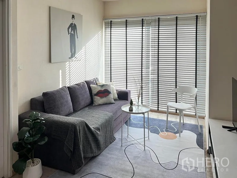 Condominium for rent in Khlong Toei, Bangkok - Bright lounge with grey sofa, blinds and small coffee table on a patterned rug.