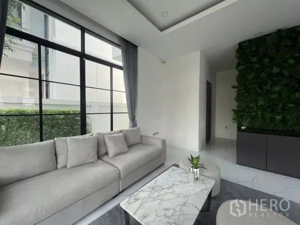 Detached House for rent in Lat Phrao, Bangkok - Contemporary lounge with grey sofa, marble coffee table and floor-to-ceiling black-framed windows beside a vertical garden wall.
