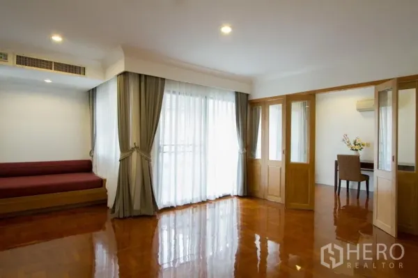 Apartment for rent in Watthana, Bangkok - Bright sitting area with daybed, floor‑to‑ceiling curtains, and glass doors leading to a study nook.