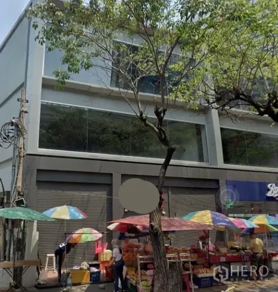 Retail Space for rent in Watthana, Bangkok - Street-facing modern retail building with large glass windows, roll-up shutters, and colorful market umbrellas under trees in Watthana, Bangkok.