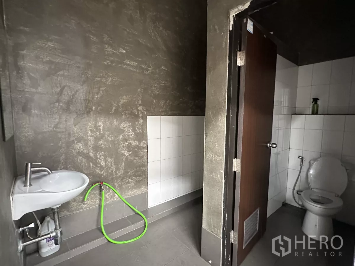 Warehouse for rent in Khlong Toei, Bangkok - Warehouse restroom featuring tiled WC, wall-mounted sink and hose against textured gray walls.