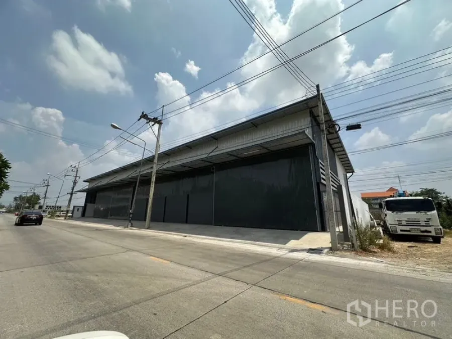 Warehouse for rent in Lam Luk Ka, Pathum Thani - Corner view of the street-side warehouse exterior with power lines and a loading ramp.
