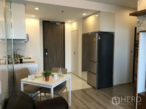 Condominium for rent in Bang Na, Bangkok - Open-plan kitchen and dining area with white table, built-in cabinets, and a stainless refrigerator by the entry.