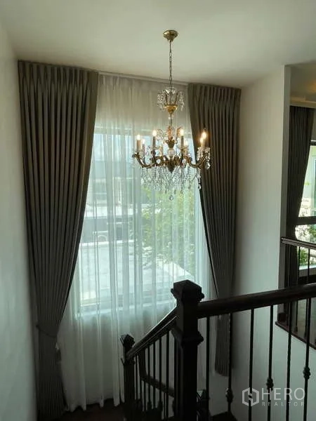 Detached House for rent or sale in Lat Krabang, Bangkok - Stairwell featuring a large curtained window, crystal chandelier and wooden banister.