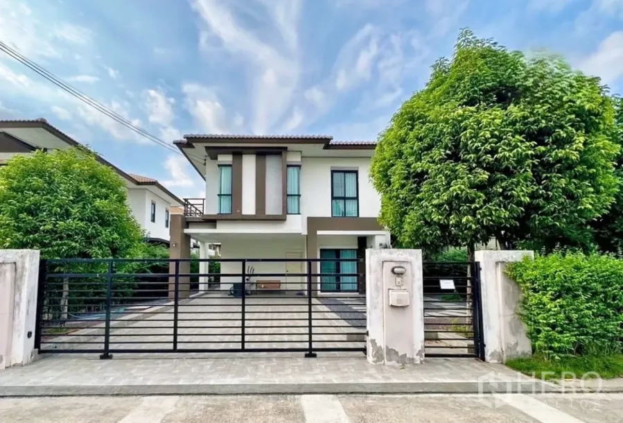Detached House for sale in Lam Luk Ka, Pathum Thani - Modern two-storey detached house with wide driveway, black metal gate and mature trees under a blue sky.