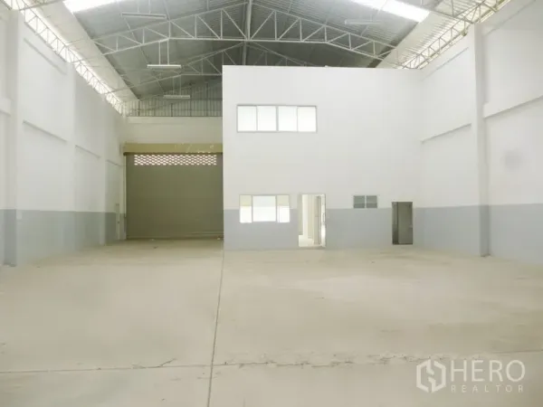 Factory for rent or sale in Pathum Thani - Spacious factory interior featuring a high steel truss roof, large roller door and a white-walled office block.