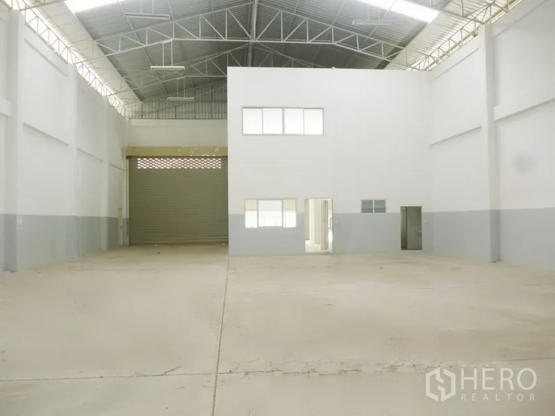 Factory for rent or sale in Pathum Thani - Spacious factory interior featuring a high steel truss roof, large roller door and a white-walled office block.