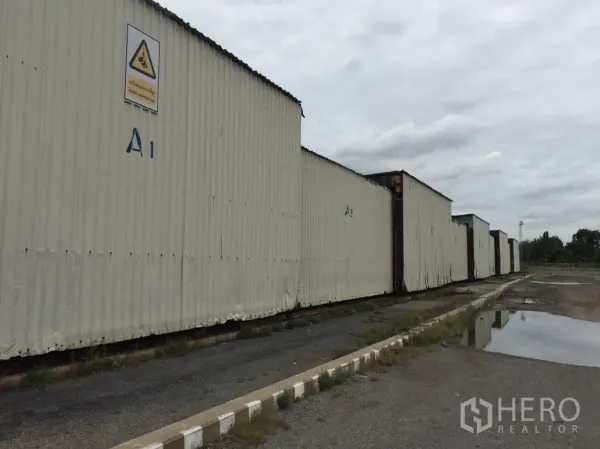 Warehouse for rent in Mueang Pathum Thani, Pathum Thani - Row of corrugated metal warehouse blocks labeled A1–A2 with wide driveway and drainage under cloudy sky.