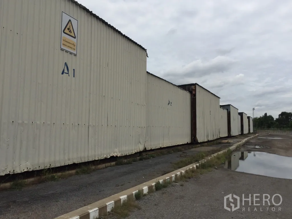 Warehouse for rent in Mueang Pathum Thani, Pathum Thani - Row of corrugated metal warehouse blocks labeled A1–A2 with wide driveway and drainage under cloudy sky.