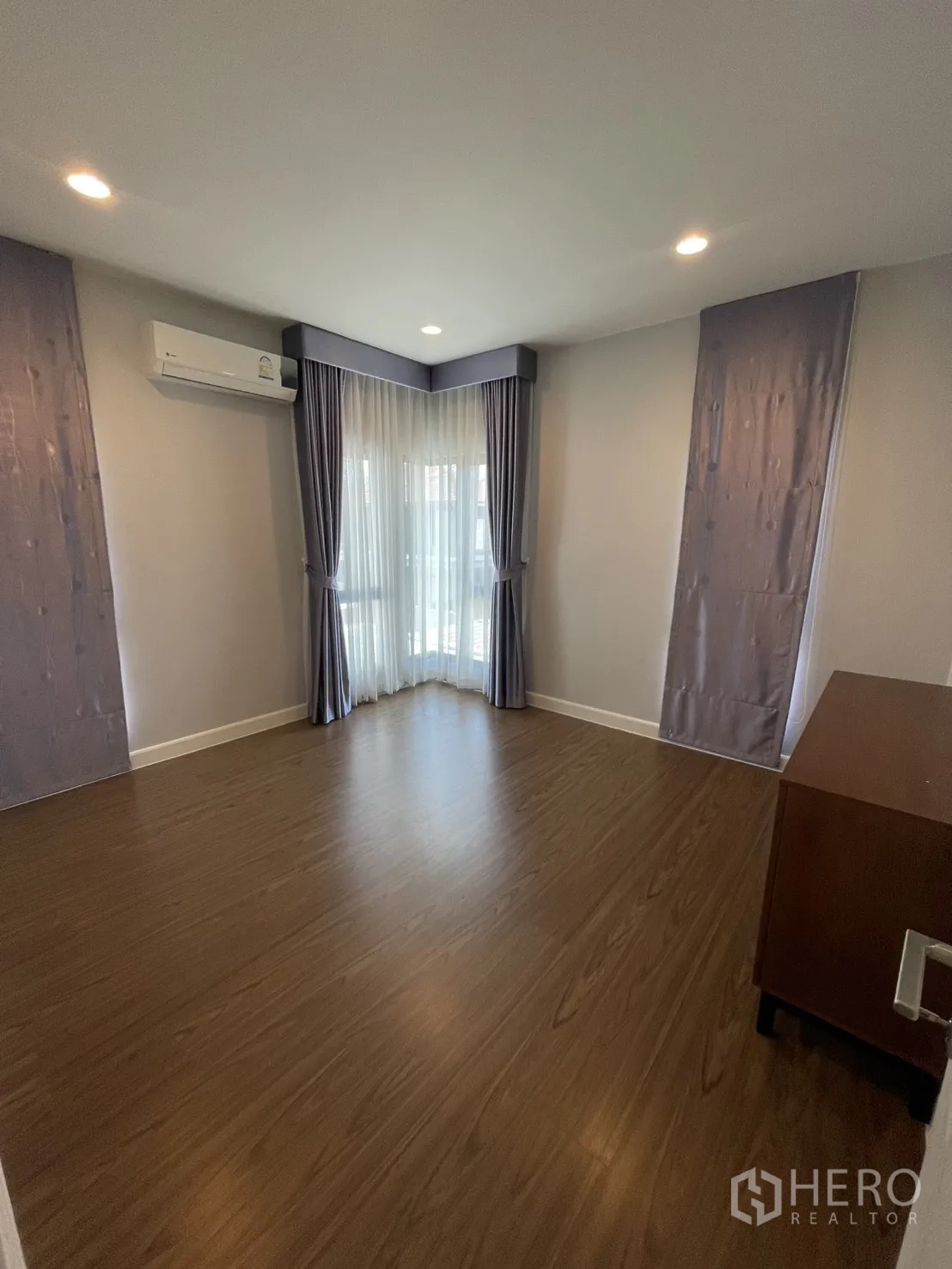 Detached House for rent or sale in Lam Luk Ka, Pathum Thani - Empty bedroom with corner windows, blackout and sheer curtains, air-conditioner and wood floors.