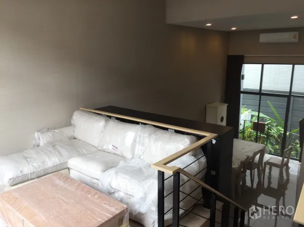 Townhouse for rent in Suan Luang, Bangkok - Mezzanine lounge with white sectional sofa, high ceiling, recessed lights and tall windows to greenery.