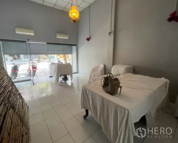 店屋 出租在Watthana, Bangkok - 一层店面空间，落地玻璃门、挑高天花与瓷砖地面，家具覆盖布套。