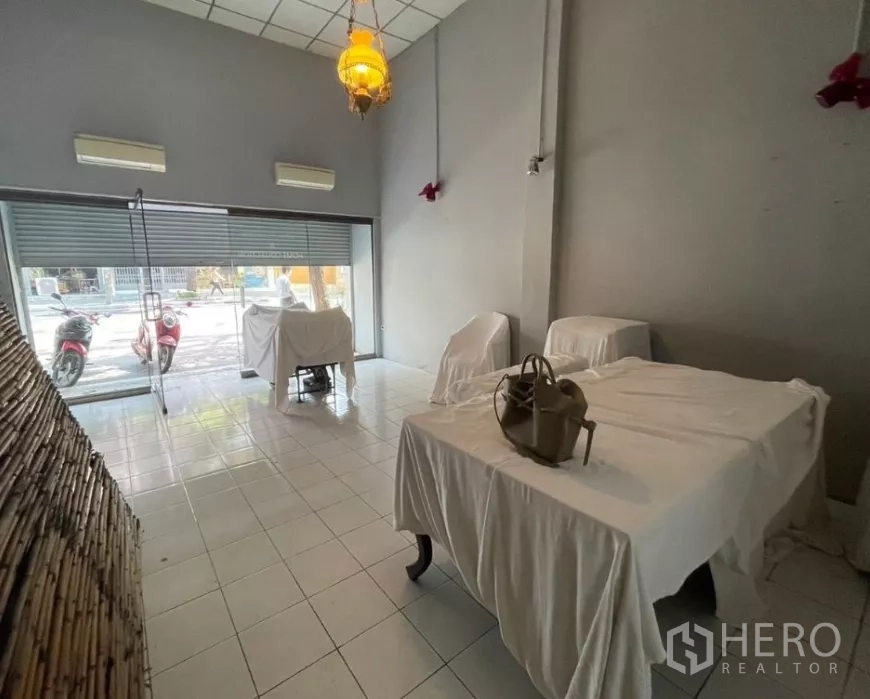 店屋 出租在Watthana, Bangkok - 一层店面空间，落地玻璃门、挑高天花与瓷砖地面，家具覆盖布套。