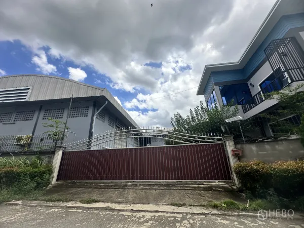 Factory for sale in Pak Kret, Nonthaburi - Gated industrial compound with a metal warehouse and a modern two‑storey office or house beside a tall fence under a cloudy sky.