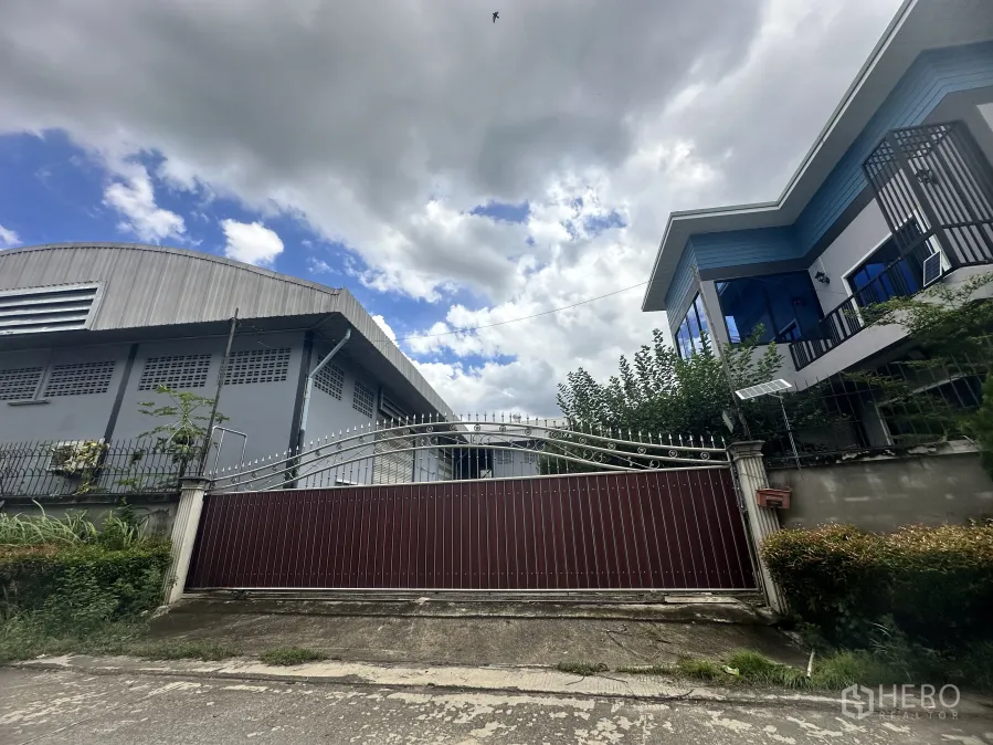 Factory for sale in Pak Kret, Nonthaburi - Gated industrial compound with a metal warehouse and a modern two‑storey office or house beside a tall fence under a cloudy sky.