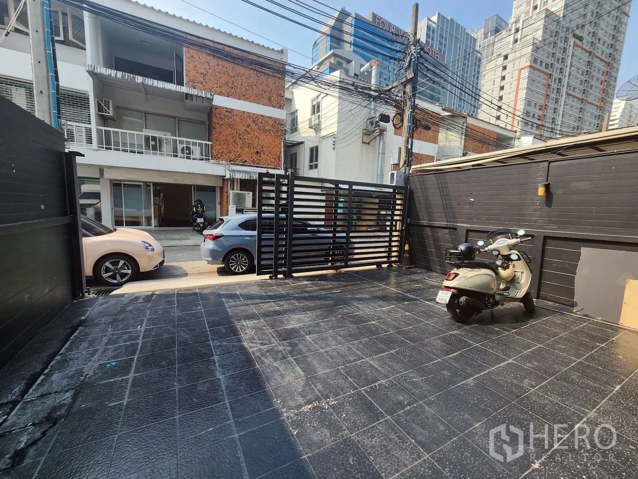 家庭办公室 for rent in Khlong Toei, Bangkok - 带大门的铺装前院和踏板车，正对低层建筑与城市高楼。