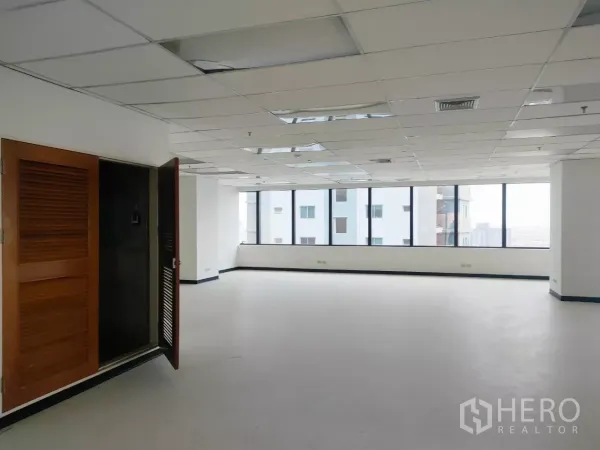 Office Space for rent in Huai Khwang, Bangkok - Vacant office interior featuring long window walls, a drop ceiling, and a wooden utility cabinet by the entrance.