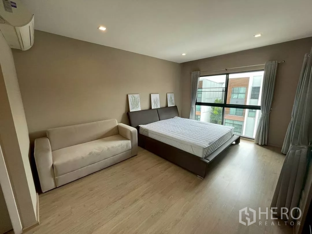 Townhouse for rent in Suan Luang, Bangkok - Master bedroom with sofa seating, large bed and wide windows facing neighboring buildings.