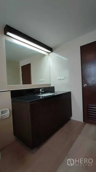 Condominium for rent in Khlong Toei, Bangkok - Vanity area with long backlit mirror and dark countertop in the condo bathroom.